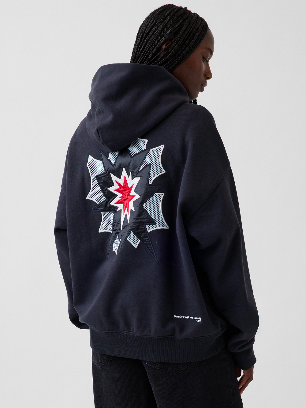 A model shown from the back wearing a black hoodie with a large graphic print inspired by Roy Lichtenstein's work Standing Explosion on the back