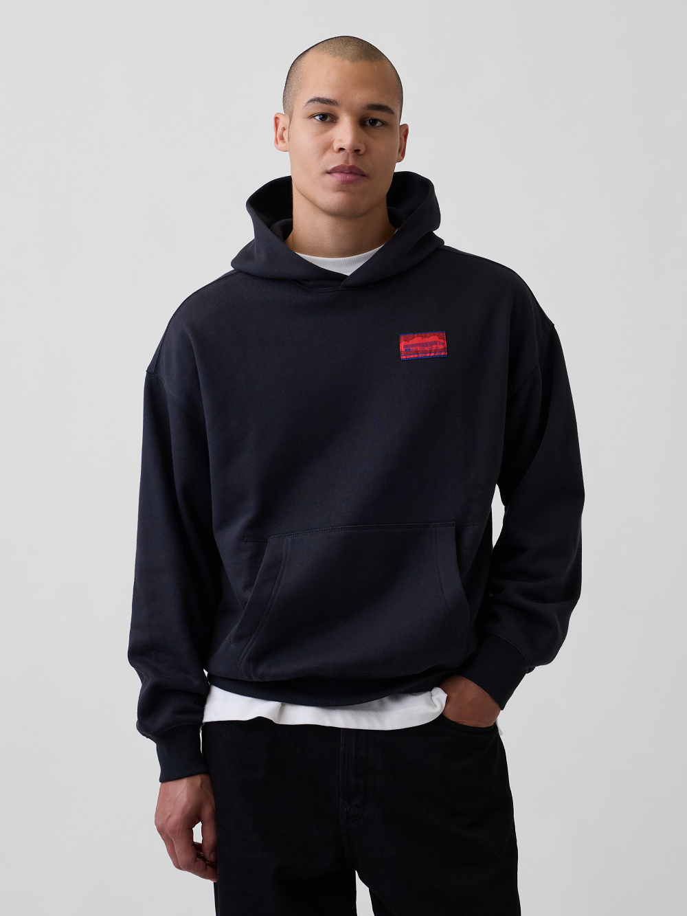 A model wearing a black pullover hoodie with a red and blue woven label on the left chest, layered over a white t-shirt and black pants, photographed against a neutral light gray background