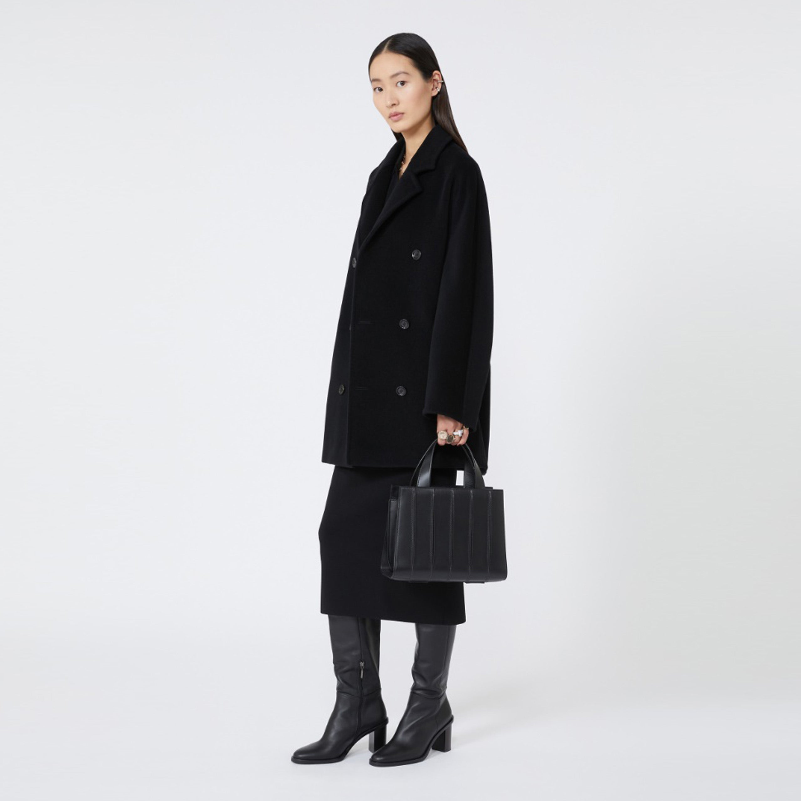 Model wearing a black coat and holding a black handbag on a white background