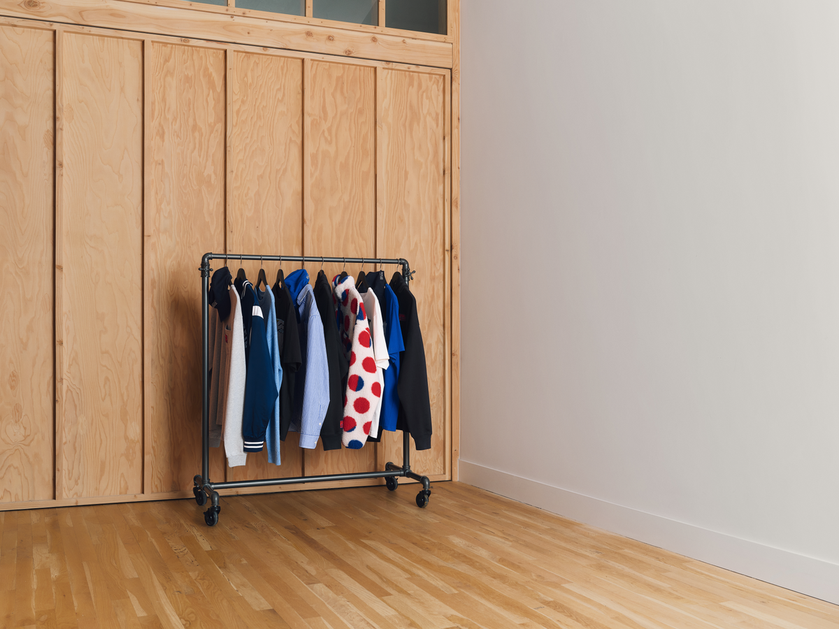 A black metal rolling clothing rack with wheels, holding a selection of of garments and accessories from the Gap – Lichtenstein – Whitney collaboration set against a light wood paneled wall in a minimalist room